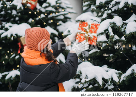 Back view of a woman decorating a snow-covered Christmas tree with wrapped gift boxes. Holiday decor trends and festive outdoor winter scenes Back view of a woman decorating a snow-covered Christmas tree with wrapped gift boxes. Holiday decor trends and festive outdoor winter scenes 119267717
