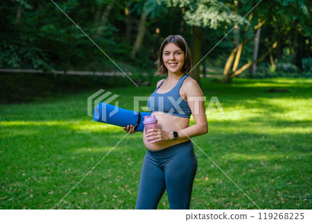 Pregnant woman walking in park with sports bottle and yoga mat in hands 119268225