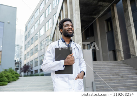 Man doctor with beard holding clipboard with medical documents posing near modern clinic 119268254