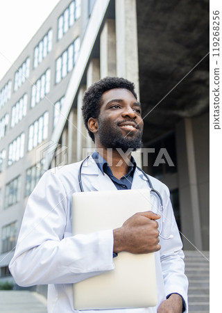 Smiling doctor holding laptop in hands standing outdoor modern hospital 119268256