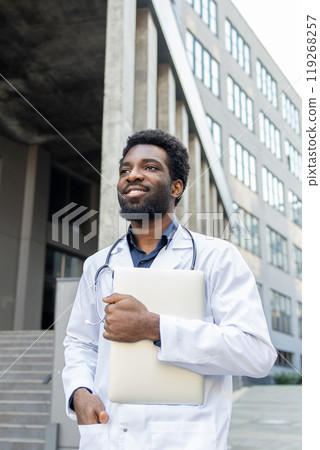 Doctor standing outdoors clinic holding laptop in one hand ready to connect with patients online Doctor standing outdoors clinic holding laptop in one hand ready to connect with patients online 119268257