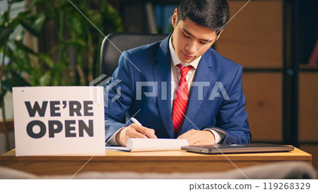 Businessman sitting at table with we are open sign and signing documents. Contract signing Businessman sitting at table with we are open sign and signing documents. Contract signing 119268329