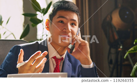 Young businessman sitting at desk in his office and leading conversation via mobile phone with clients and partners. 119268355