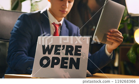 Focused Asian man sitting at desk in his office, making notes, holding tablet. Conversation with partners and clients. We are open sign on foreground 119268366