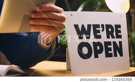 Businessman in formal wear sitting at desk with we are open sign, holding tablet and making notes. Cropped image 119268369