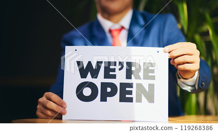 Cropped image of man in formal wear sitting in with we are open sign in hands, meaning opening new business, promotion of new ventures. Cropped image of man in formal wear sitting in with we are open sign in hands, meaning opening new business, promotion of new ventures. 119268386