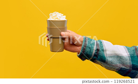 Woman's hand holds mug of Bombardino, garnished with whipped cream, against warm yellow studio background. Woman's hand holds mug of Bombardino, garnished with whipped cream, against warm yellow studio background. 119268521