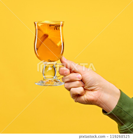 Woman's hand holds glass of Hot apple cider garnished with citrus slice and cinnamon stick, against warm yellow studio background. Woman's hand holds glass of Hot apple cider garnished with citrus slice and cinnamon stick, against warm yellow studio background. 119268525