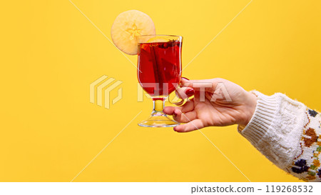 Woman's hand holds a glass of mulled wine, garnished with apple slice and cinnamon stick, against warm yellow studio background. Mulled wine. 119268532