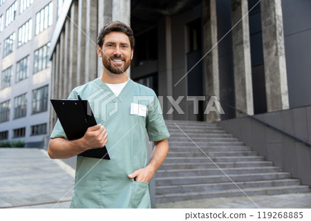 Man doctor with beard holding clipboard with medical documents posing near modern clinic Man doctor with beard holding clipboard with medical documents posing near modern clinic 119268885
