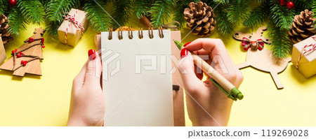 Top view of female hand making some notes in noteebok on yellow background. New Year decorations and toys. Christmas time concept Top view of female hand making some notes in noteebok on yellow background. New Year decorations and toys. Christmas time concept 119269028