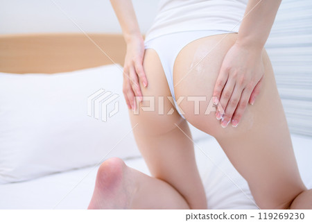 一位穿著內衣的年輕苗條女人在床上塗抹身體霜。 119269230