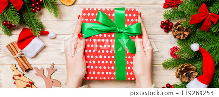 Top view of a woman holding a gift box in her hands on festive wooden background. Fir tree and Christmas decorations. New year time concept Top view of a woman holding a gift box in her hands on festive wooden background. Fir tree and Christmas decorations. New year time concept 119269253
