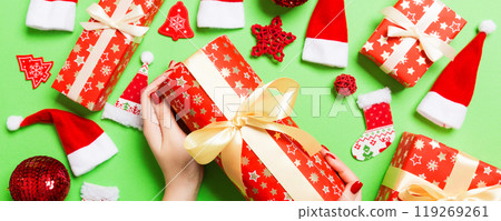 Top view of female hands holding a Christmas present on festive green background. Santa hats and holiday decorations. New Year holiday concept Top view of female hands holding a Christmas present on festive green background. Santa hats and holiday decorations. New Year holiday concept 119269261