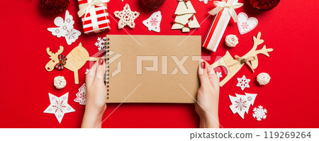 Top view of female hand making some notes in noteebok on colorful background. New Year decorations and toys. Christmas time concept Top view of female hand making some notes in noteebok on colorful background. New Year decorations and toys. Christmas time concept 119269264