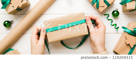Woman is packing surprise gift for Christmas holidays, sitting at workplace with craft tools, top view. Woman wrapping Christmas gift at colored table Woman is packing surprise gift for Christmas holidays, sitting at workplace with craft tools, top view. Woman wrapping Christmas gift at colored table 119269315