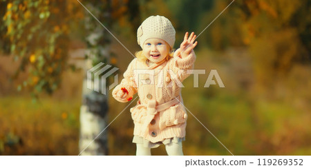 Happy little cheerful child having fun playing in autumn park in warm knitted sweater and hat 119269352