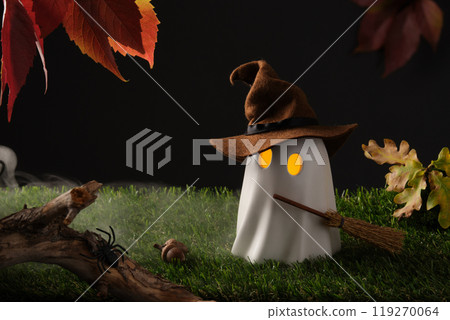 Cute Halloween ghost with witch hat and broom, glowing eyes on green grass with autumn leaves and spider, spooky autumn scene Cute Halloween ghost with witch hat and broom, glowing eyes on green grass with autumn leaves and spider, spooky autumn scene 119270064