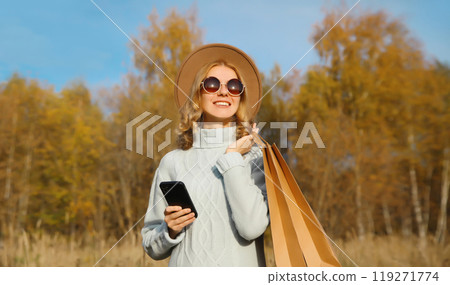 Stylish happy young woman with shopping bag, smartphone in autumn park, girl smiling using phone 119271774