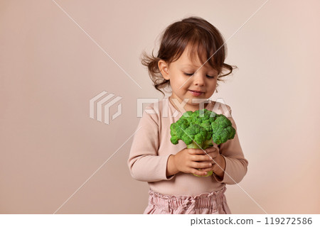 smiling child girl looking at fresh green raw broccoli on beige background. healthy baby food. kid loves vegetables smiling child girl looking at fresh green raw broccoli on beige background. healthy baby food. kid loves vegetables 119272586