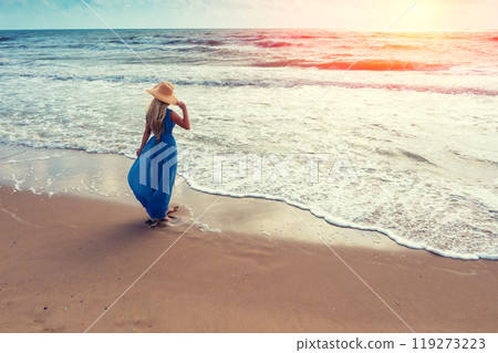 Seascape during sunrise. Woman on the beach. Young happy woman in a long fluttering blue dress and straw hat walks on the seashore 119273223