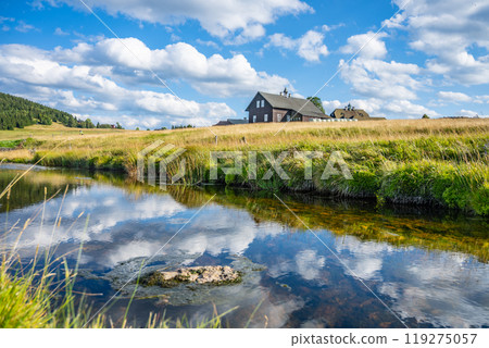 The tranquil village of Jizerka, nestled in the Jizera Mountains, showcases charming cottages beside a reflective pond. Fluffy clouds drift in the blue sky, enhancing this picturesque rural setting. The tranquil village of Jizerka, nestled in the Jizera Mountains, showcases charming cottages beside a reflective pond. Fluffy clouds drift in the blue sky, enhancing this picturesque rural setting. 119275057