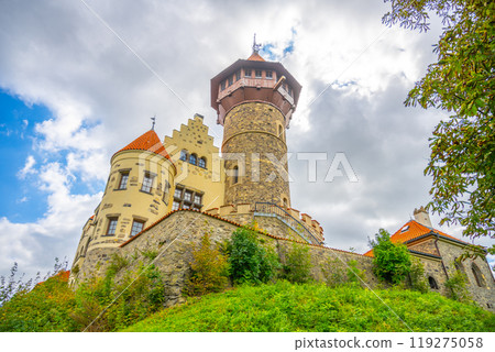 The lookout tower of Hnevin Castle stands majestically amid lush greenery and dynamic clouds, showcasing its unique architectural style in Most, Czechia on a picturesque day. 119275058