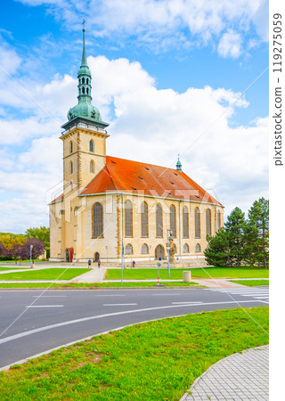 The Church of the Assumption of the Virgin Mary in Most showcases its stunning late Gothic architecture under a partly cloudy sky. Visitors stroll around the lush green surroundings. The Church of the Assumption of the Virgin Mary in Most showcases its stunning late Gothic architecture under a partly cloudy sky. Visitors stroll around the lush green surroundings. 119275059