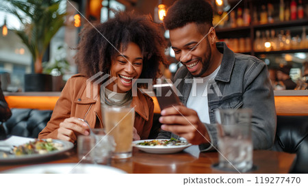 Couple enjoying dinner at a cozy restaurant while looking at a smartphone, sharing smiles and laughter 119277470