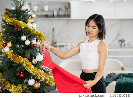 Sad young Asian girl trying to choose a dress standing next to Xmas tree 119281018