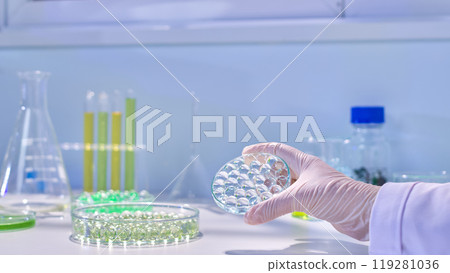Advertising photo in modern laboratory from close up front view, a petri dish of clear gel balls being held in scientist's hand, surrounded by variety experiment instruments 119281036