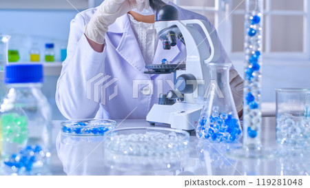 The experiment scene with blue and white hydrogel, a scientist is sitting at the lab table and looking into microscope to observe a blue gel ball. Front view, minimal laboratory background The experiment scene with blue and white hydrogel, a scientist is sitting at the lab table and looking into microscope to observe a blue gel ball. Front view, minimal laboratory background 119281048