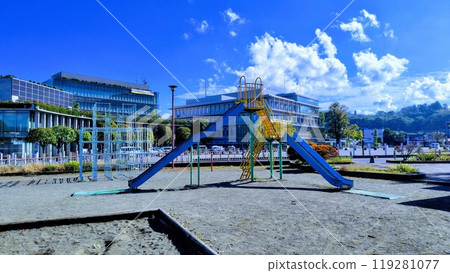 Sandbox, slide and jungle gym in a park, with buildings and blue sky and clouds in the background Sandbox, slide and jungle gym in a park, with buildings and blue sky and clouds in the background 119281077