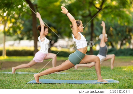 Skilled happy young female teacher demonstrating variation of warrior pose of yoga for group women at city park 119281084