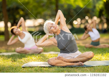 Happy old lady and class of calm sports women in activewear stretching muscles and doing tilts while sitting in lotus pose on mats during yoga training together at city park 119281102