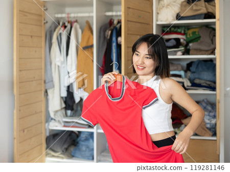 Happy young Asian girl trying to choose a dress standing by wardrobe 119281146