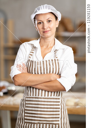 Confident young female bakery in apron posing with arms crossed indoors in bakery kitchen 119281150
