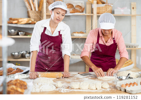Two skilled female bakers in apron holding rolling pin in hands and rolling out dough in bakery kitchen Two skilled female bakers in apron holding rolling pin in hands and rolling out dough in bakery kitchen 119281152