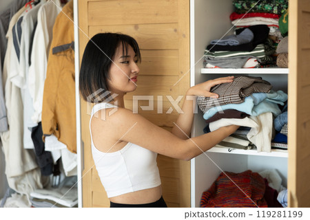 Happy young Asian girl putting some items of clothing in the wardrobe Happy young Asian girl putting some items of clothing in the wardrobe 119281199