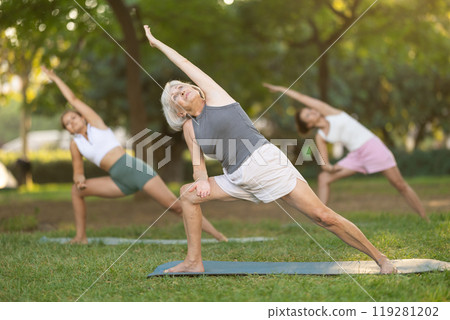 Sporty energetic mature female teacher doing exercises on pilates mat and demonstrating variation of warrior pose of yoga for group women at city park. Fitness concept Sporty energetic mature female teacher doing exercises on pilates mat and demonstrating variation of warrior pose of yoga for group women at city park. Fitness concept 119281202