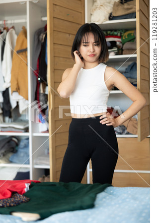 Sad young Asian girl trying to choose a dress standing by wardrobe 119281203