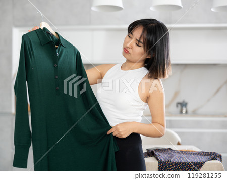 Sad young Asian girl trying to choose a dress in the kitchen 119281255