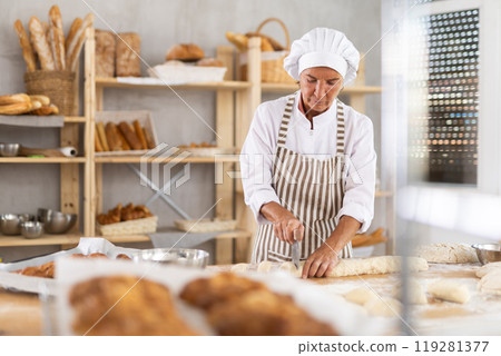 Mature woman baker cuts dough in bakehouse Mature woman baker cuts dough in bakehouse 119281377