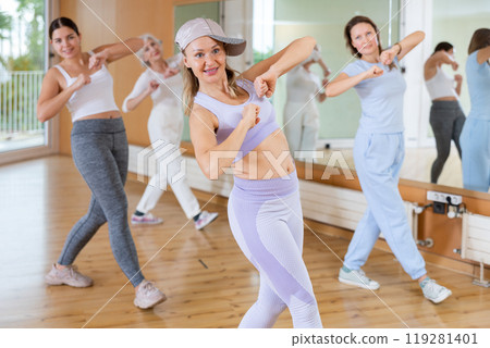 Joyful young mature women in sportswear learning new movements with hands at chest level while dancing together in hall 119281401