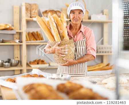Happy mature female baker posing with basket of freshly baked baguettes in bakery, demonstrating concept of food industry and baker profession 119281463