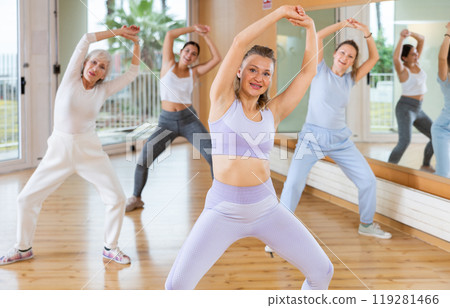 Group of young mature emotional female dancers raising both hands and having energetic training in dance hall 119281466