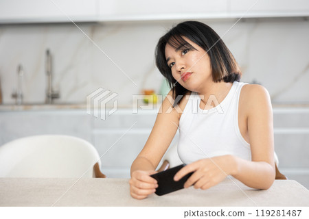 Sad young Asian girl sitting at the kitchen table with smartphone in hand 119281487