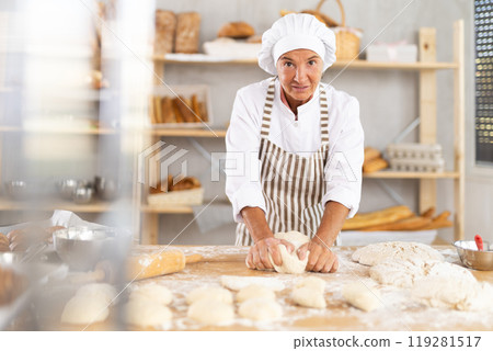 Mature woman professional baker in apron standing at work table, kneading dough and forming dough in bakery kitchen Mature woman professional baker in apron standing at work table, kneading dough and forming dough in bakery kitchen 119281517