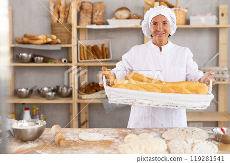 Happy old woman baker with joyful expression holds tray of freshly baked baguettes in bakery shop, showcasing concept of food industry and catering services 119281541