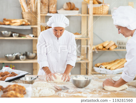 Professional mature female baker with assistant kneading dough and portioning dough in pieces during daily work in bakery Professional mature female baker with assistant kneading dough and portioning dough in pieces during daily work in bakery 119281560